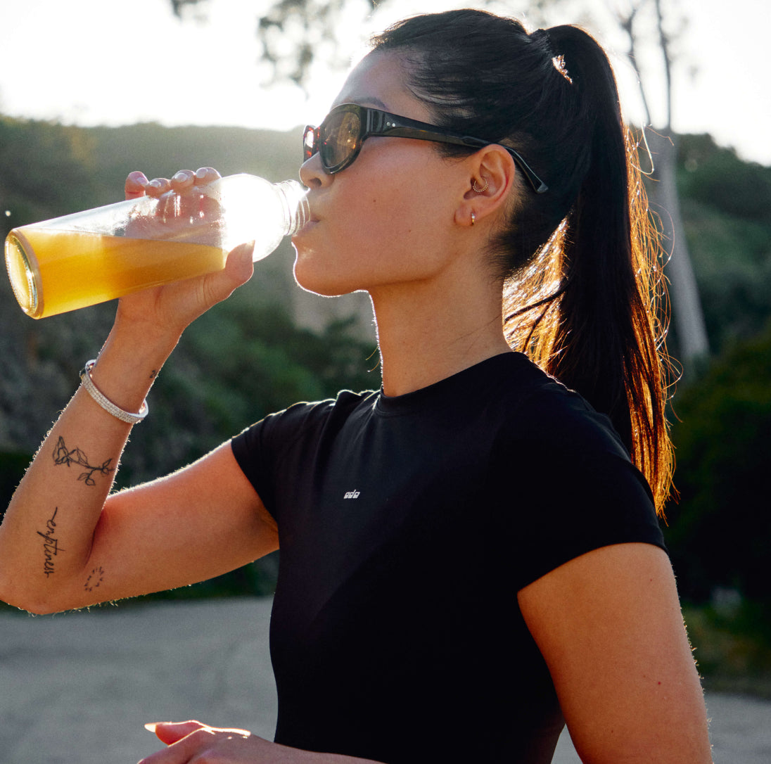 Photo of athletic woman drinking Moxie out of a glass bottle outdoors. Heightened focus and mood.